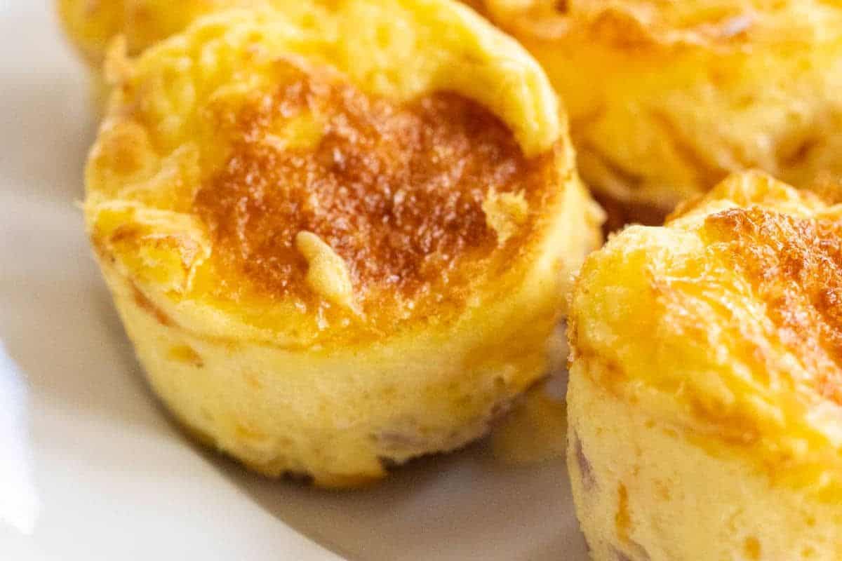 Close-up image of three mini quiches with a golden-brown crust on top, served on a white plate. The quiches appear to be fluffy and moist, with a slightly crispy texture on the surface.