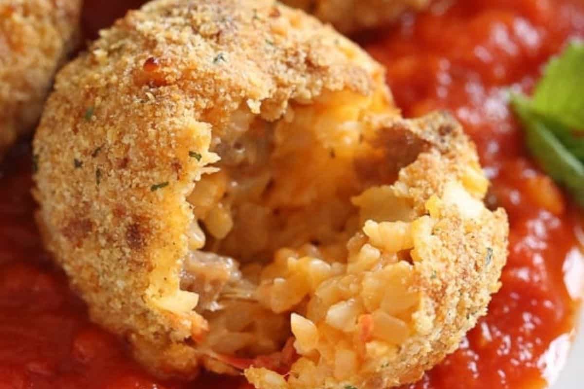 Close-up of a flavor-packed breaded rice ball with a bite taken out, revealing a cheesy filling, served on a bed of tomato sauce.