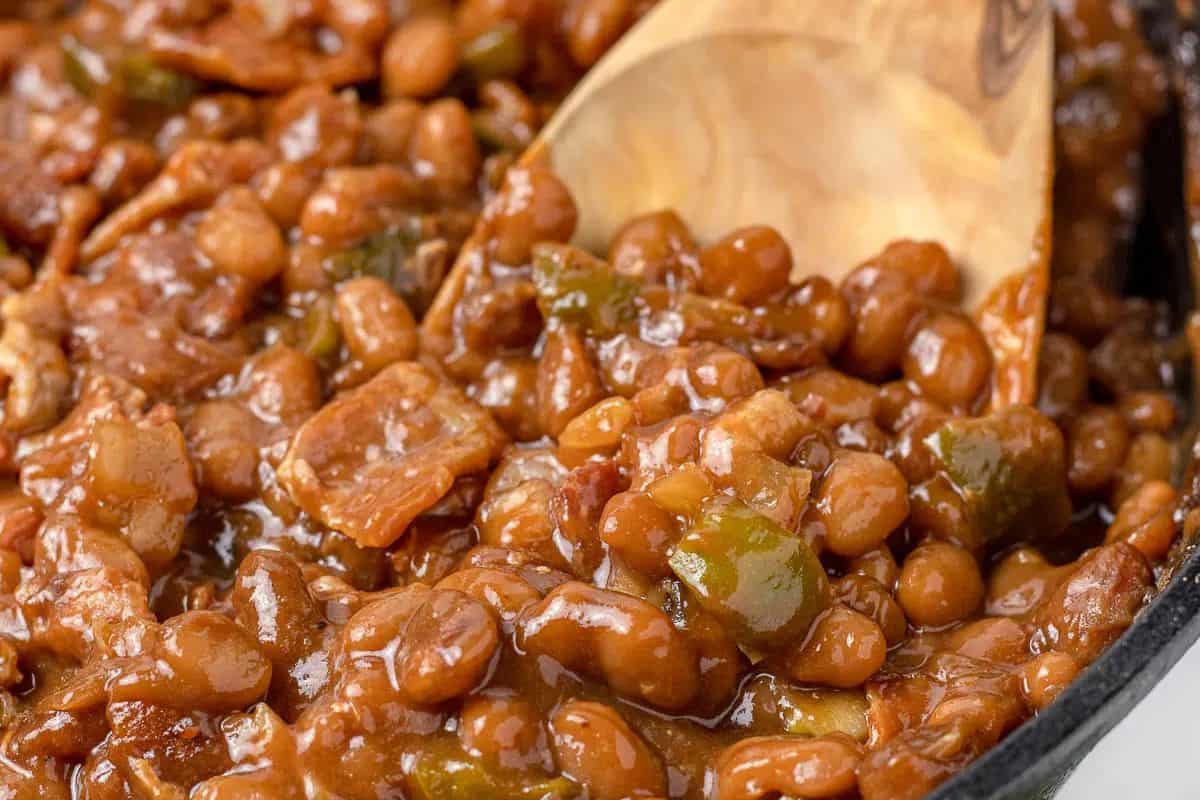 Close-up of baked beans cooked with bacon and green bell peppers in a thick, glossy sauce, being stirred with a wooden spoon.
