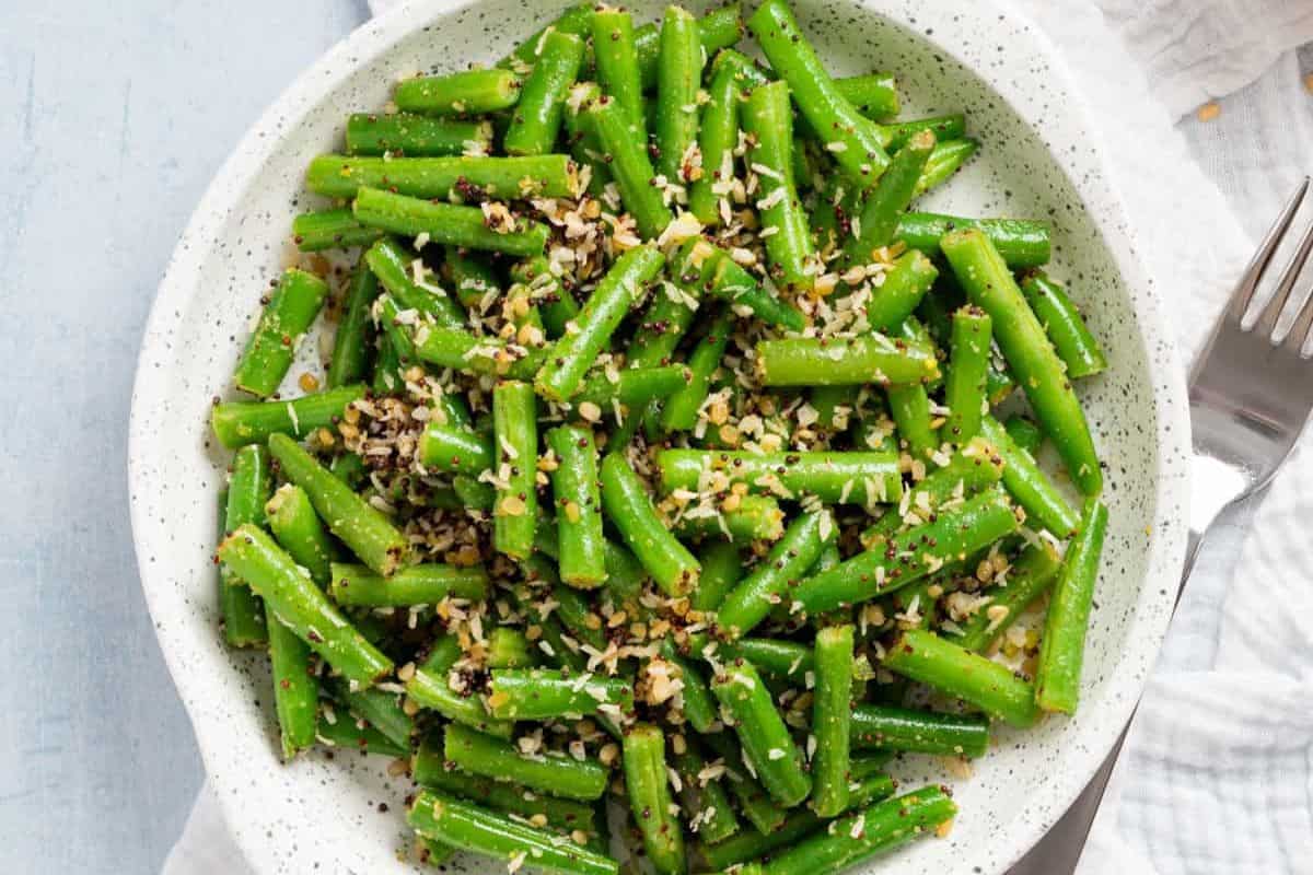 A plate of cooked green beans topped with chopped garlic and seasoning, boasting bold flavors, is placed on a white surface with a fork nearby.