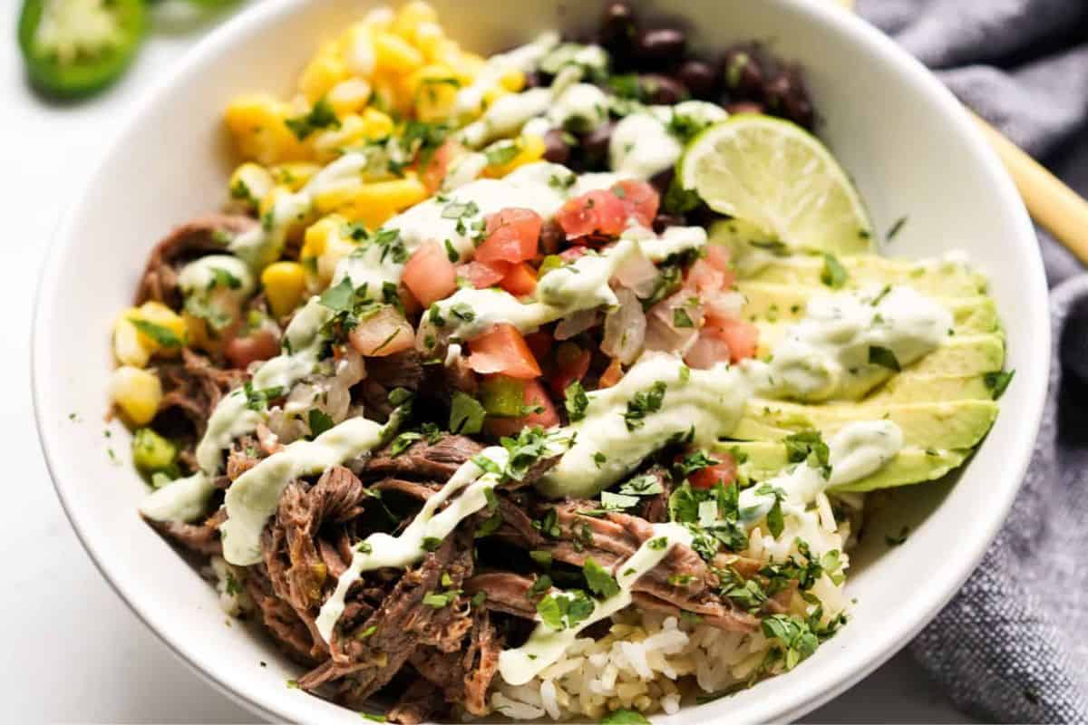 Beef burrito bowl filled with seasoned ground beef, corn, black beans, diced tomatoes, and lime wedges, served over a bed of rice.