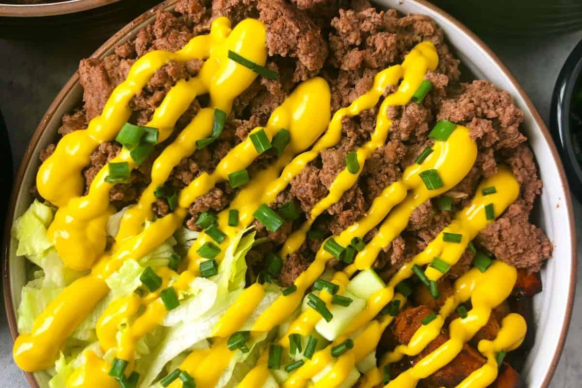 A bowl filled with chopped lettuce, cooked ground beef reminiscent of savory burgers, diced vegetables, and topped with yellow mustard and chopped green onions.