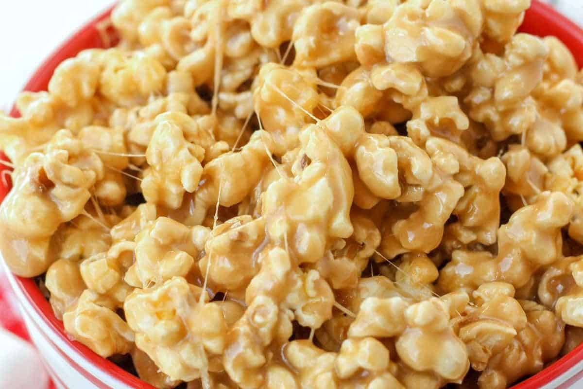 A close-up of a red and white striped bowl overflowing with caramel marshmallow popcorn, featuring sticky, stringy marshmallow strands visible among the popcorn.