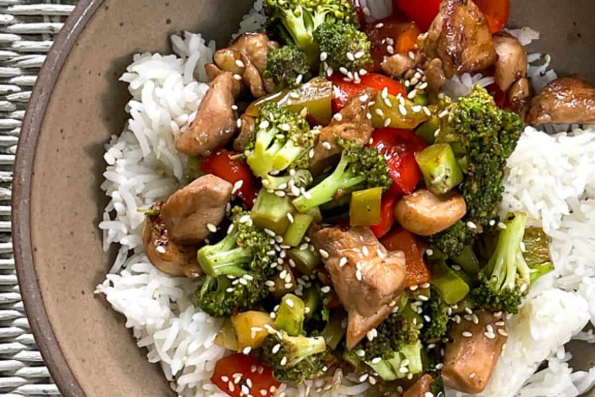 A bowl of white rice topped with colorful stir-fried vegetables, chicken, and cashews, garnished with sesame seeds.