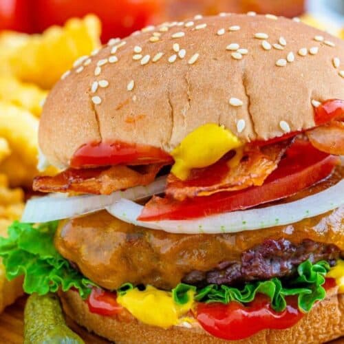 A savory cheeseburger with lettuce, tomato, onion, bacon, ketchup, and mustard in a sesame seed bun—like something from the Burger Game—served with crinkle-cut fries and pickles.