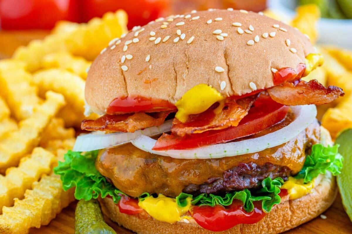 A savory cheeseburger with lettuce, tomato, onion, bacon, ketchup, and mustard in a sesame seed bun—like something from the Burger Game—served with crinkle-cut fries and pickles.