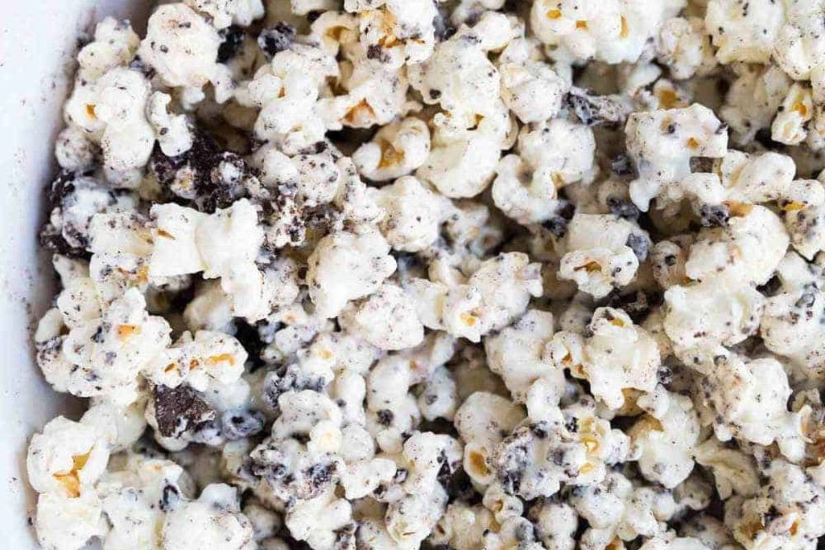  A close-up, overhead shot of a white dish filled with popcorn coated in a white creamy sauce and mixed with dark chocolate cookie crumbles, resembling cookies and cream flavor.