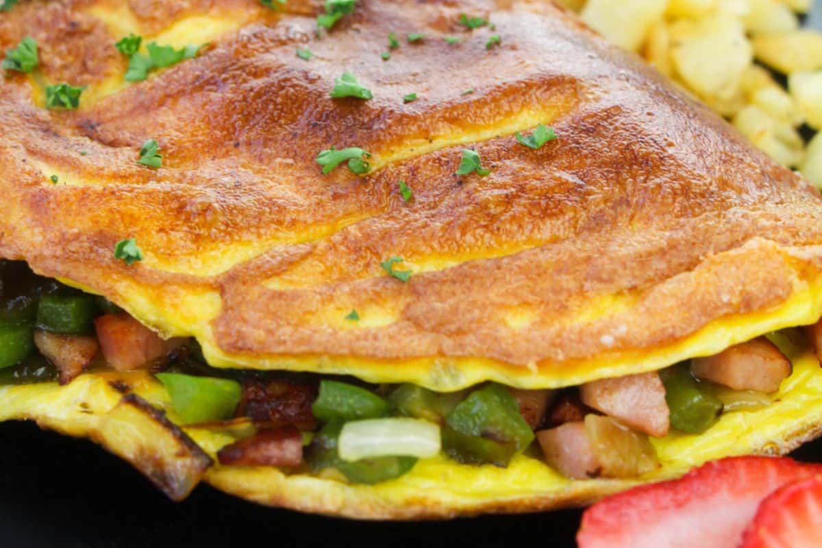 Close-up image of a golden-brown omelette garnished with small green herbs. The omelette contains visible chunks of green bell peppers, onions, and diced meat. In the background, there are diced potatoes and a slice of strawberry, adding contrast to the dish.