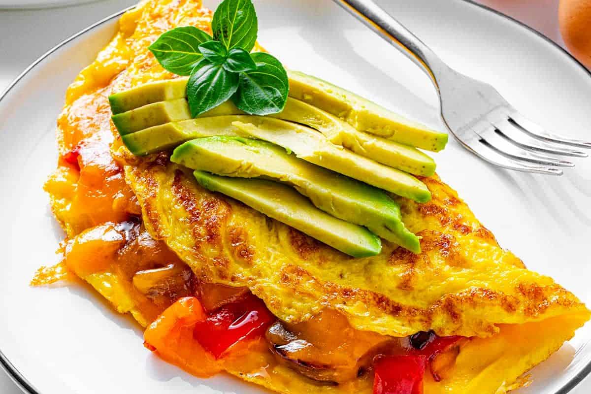 A close-up of a golden-brown omelette with a slightly crispy outer layer, filled with diced bell peppers, onions. The omelette is garnished with finely chopped parsley and avocado