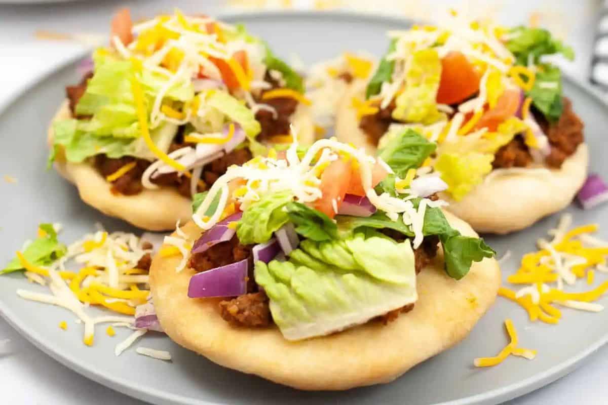 Three frybread pieces topped with seasoned ground beef, lettuce, tomato, red onion, and shredded cheese are arranged on a gray plate.