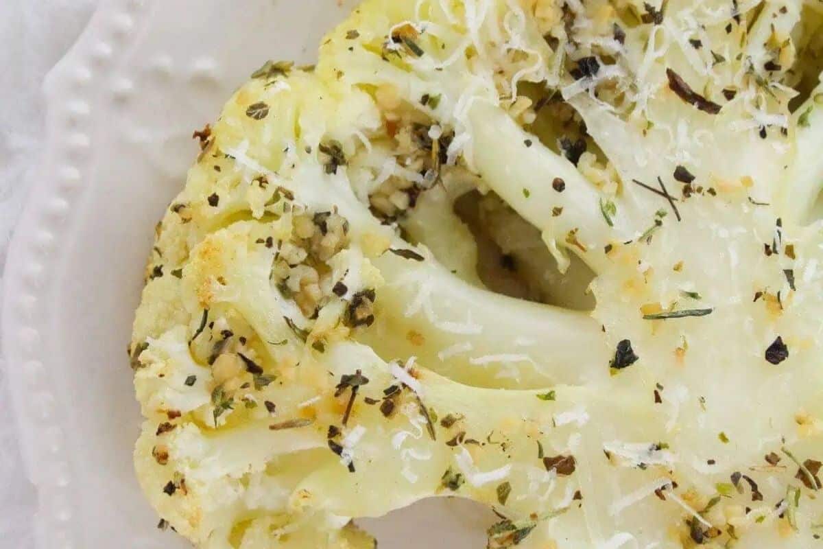 A close-up of a flavor packed cauliflower steak topped with herbs, garlic, and shredded cheese on a white plate.
