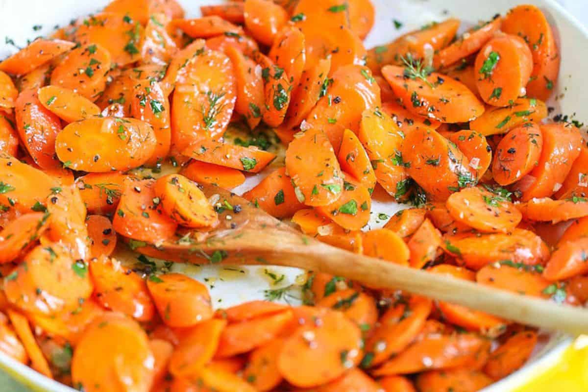 A wooden spoon resting in a pan of sliced, brightly orange carrots tossed with fresh green herbs and possibly garlic.