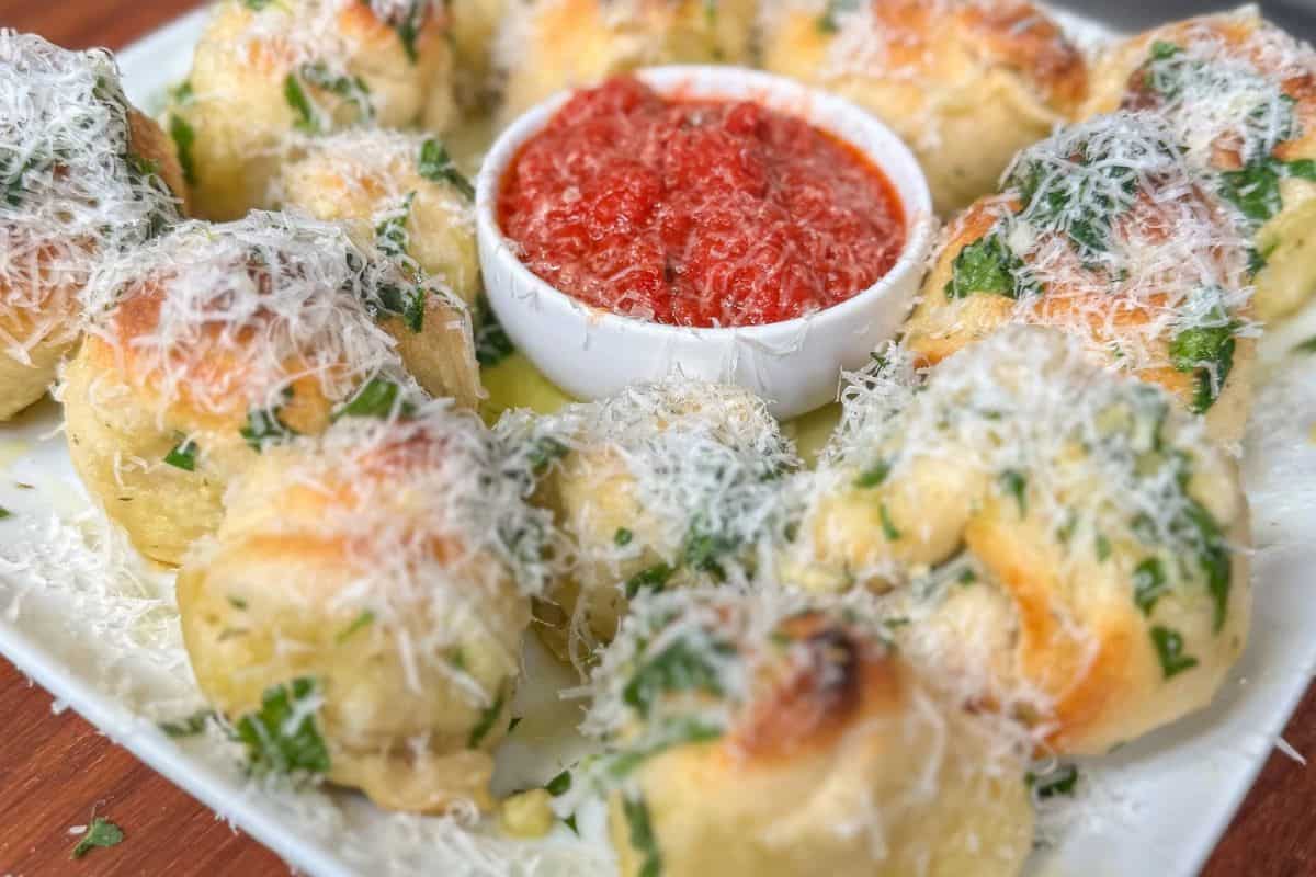 Golden garlic knots topped with cheese, served with a side of red marinara sauce.