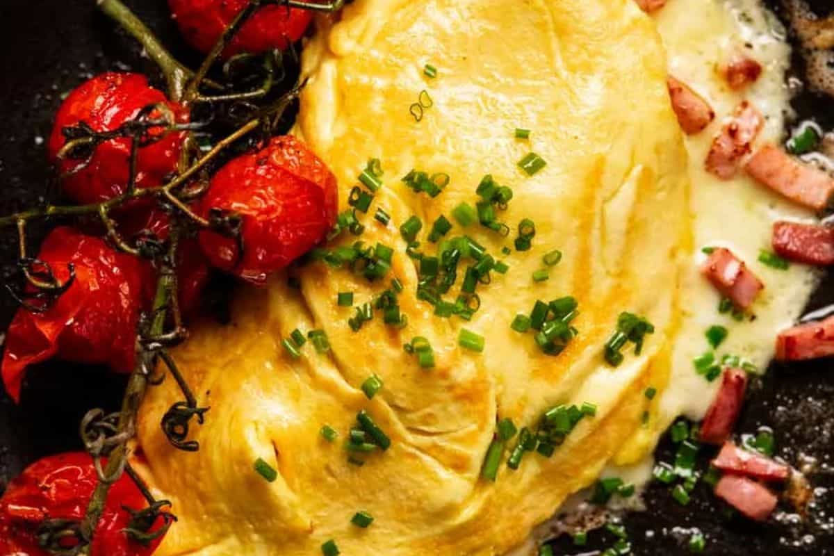 Close-up of a creamy folded omelet garnished with chopped chives, accompanied by blistered vine cherry tomatoes and diced ham melting into cheese in a skillet.