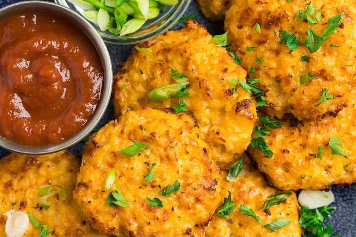 Golden-brown fritters garnished with chopped green onions and parsley, served with a side of red dipping sauce and sliced green onions.