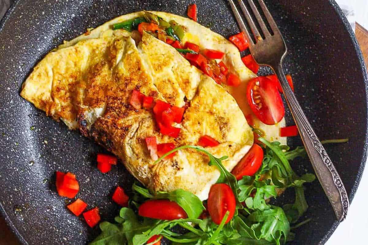 Golden-brown omelet served in a skillet, filled with sautéed vegetables and garnished with chopped tomatoes and arugula, with a fork resting beside it.