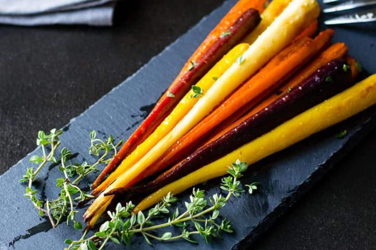 A dark slate serving platter holds several long, whole roasted carrots of various colors (orange, yellow, purple, and red), garnished with fresh thyme.