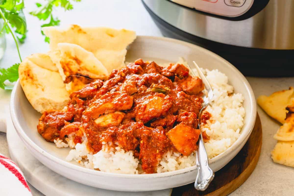 A plate of white rice topped with a red, saucy chicken curry featuring bold flavors, served with naan bread and a fork next to an Instant Pot.