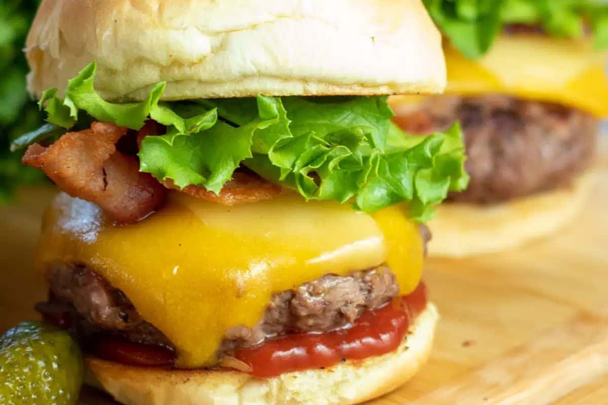 A savory burger with cheddar cheese, bacon, lettuce, and ketchup in a bun, served on a wooden surface with a pickle on the side.
