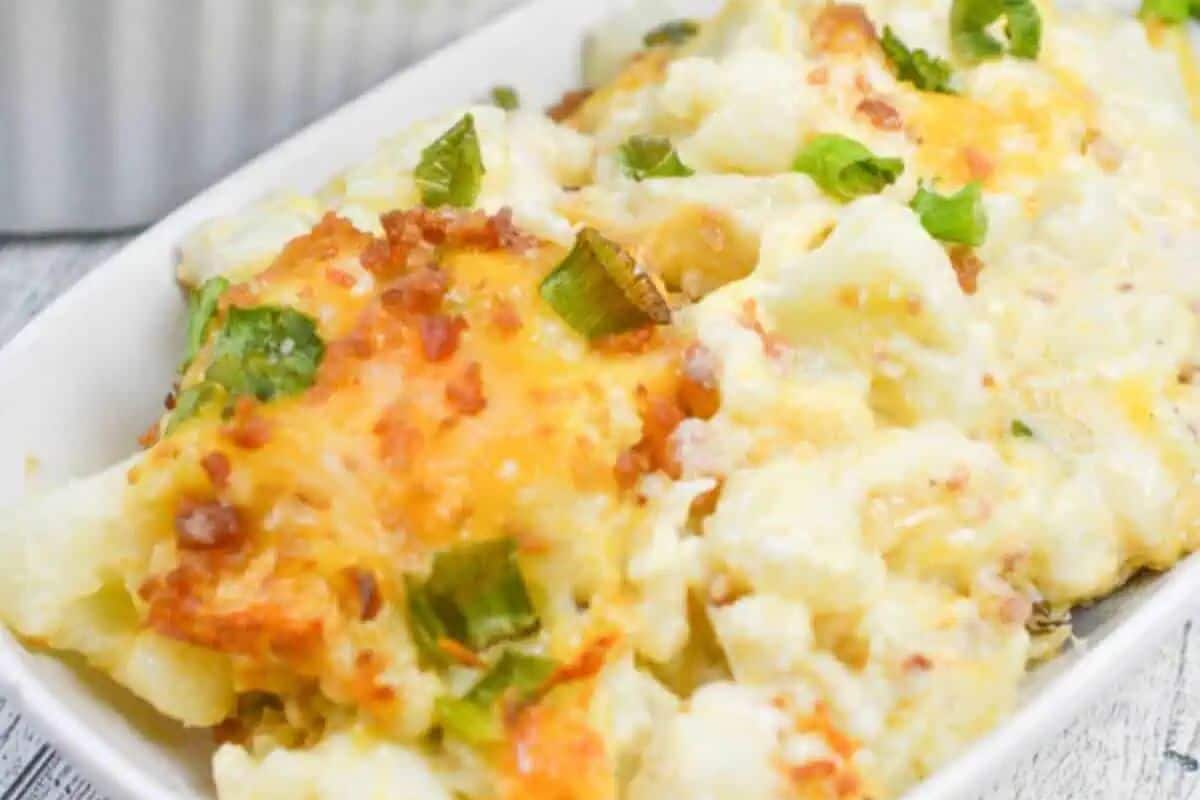A close-up of a flavor packed cauliflower casserole topped with melted cheese, chopped green onions, and bits of bacon in a white dish.