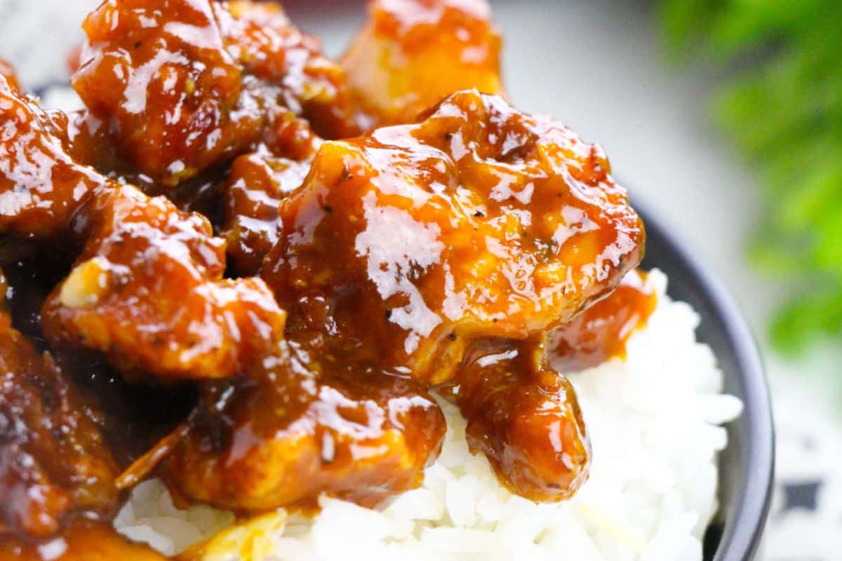 Close-up of saucy orange chicken pieces served over a bed of white rice in a bowl.