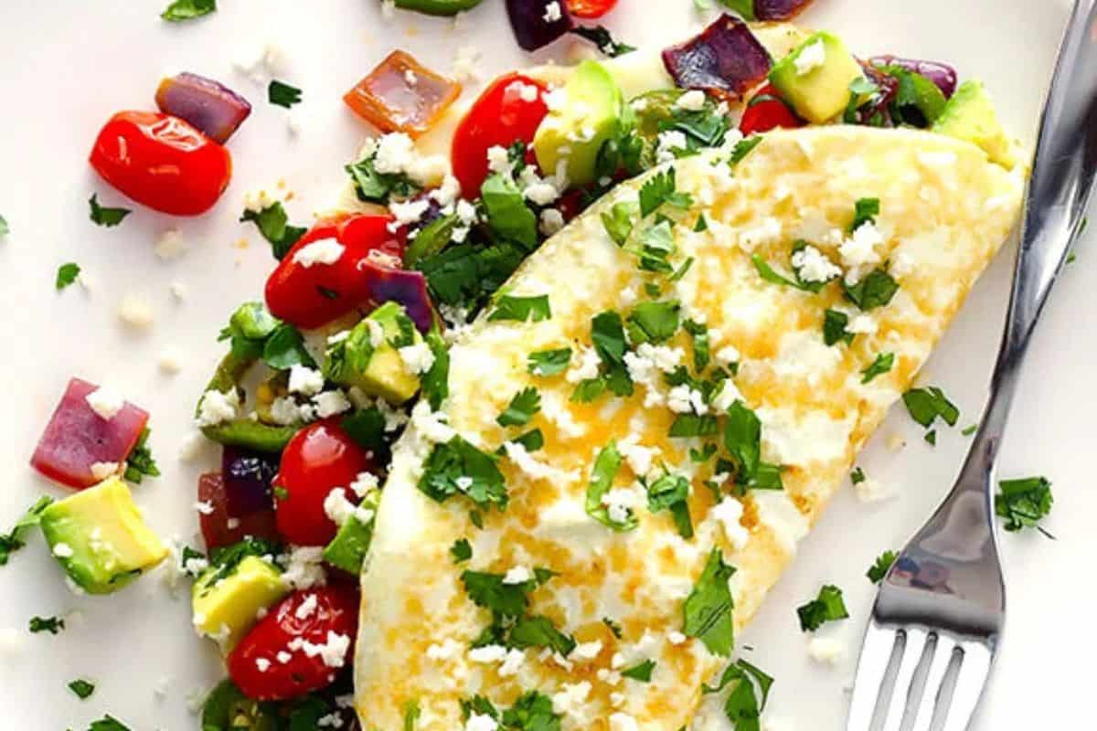 A plate with a folded omelette garnished with chopped cilantro and crumbled cheese. Next to the omelette is a colorful mix of diced vegetables including cherry tomatoes, avocado, red onion, and bell peppers. A fork is placed on the right side of the plate.