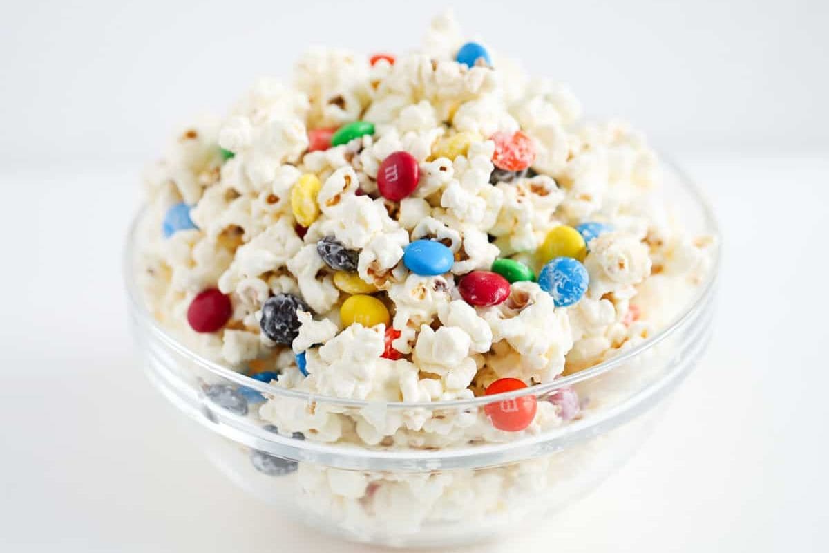 A clear glass bowl filled with white-coated popcorn mixed with colorful M&M candies, set against a white background.