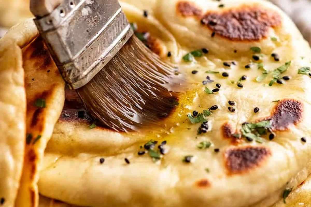 A pastry brush applies melted butter to freshly cooked naan bread topped with herbs and black sesame seeds, creating bold flavors inspired by classic Indian recipes.