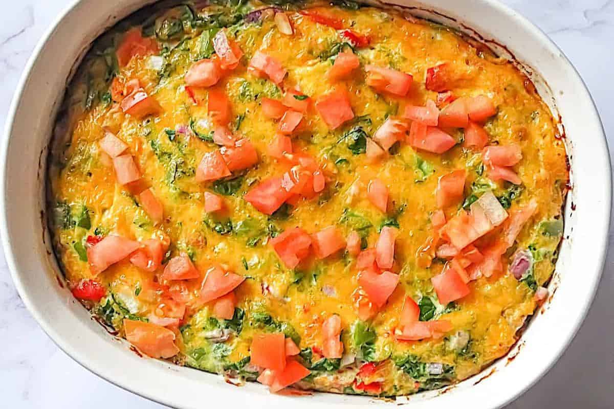 Top-down view of a colorful oven-baked omelet in a white baking dish, topped with chopped fresh tomatoes and filled with vegetables and melted cheese.