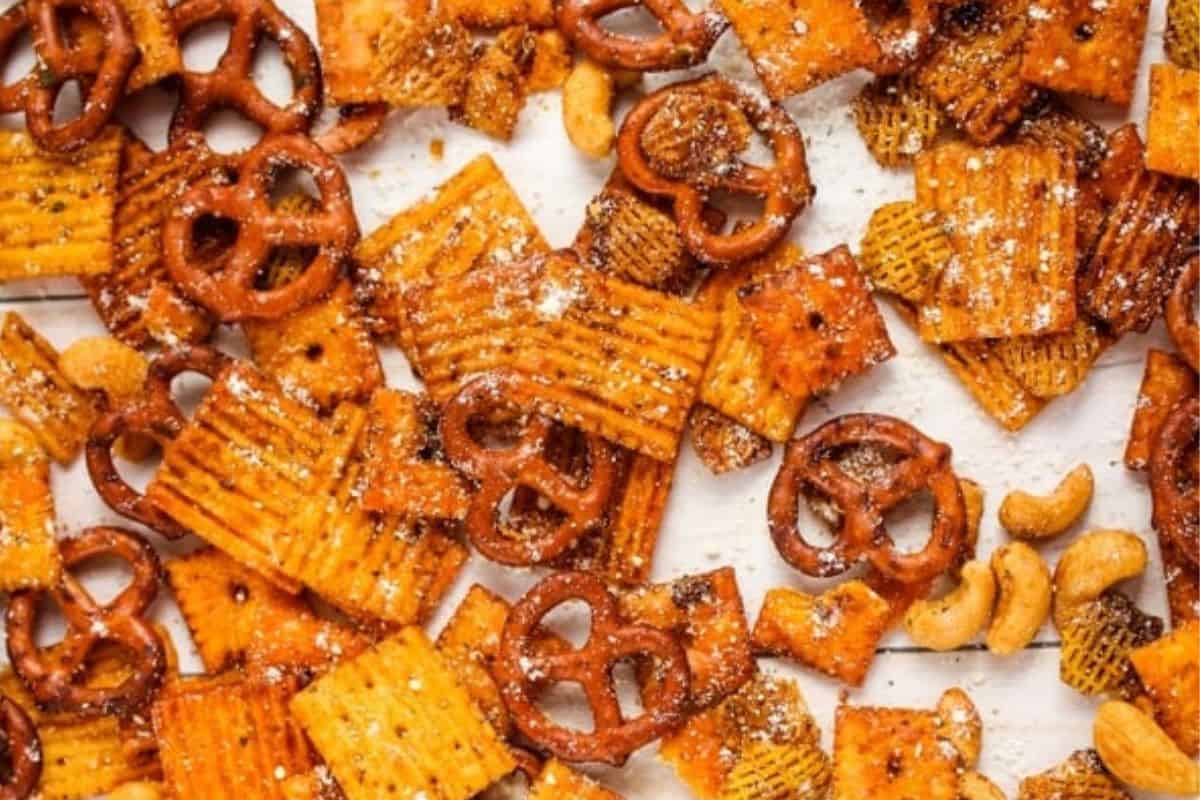 Parmesan ranch snack mix scattered on a white surface, featuring pretzels, cheese crackers, Chex cereal, and cashews coated in a savory seasoning.