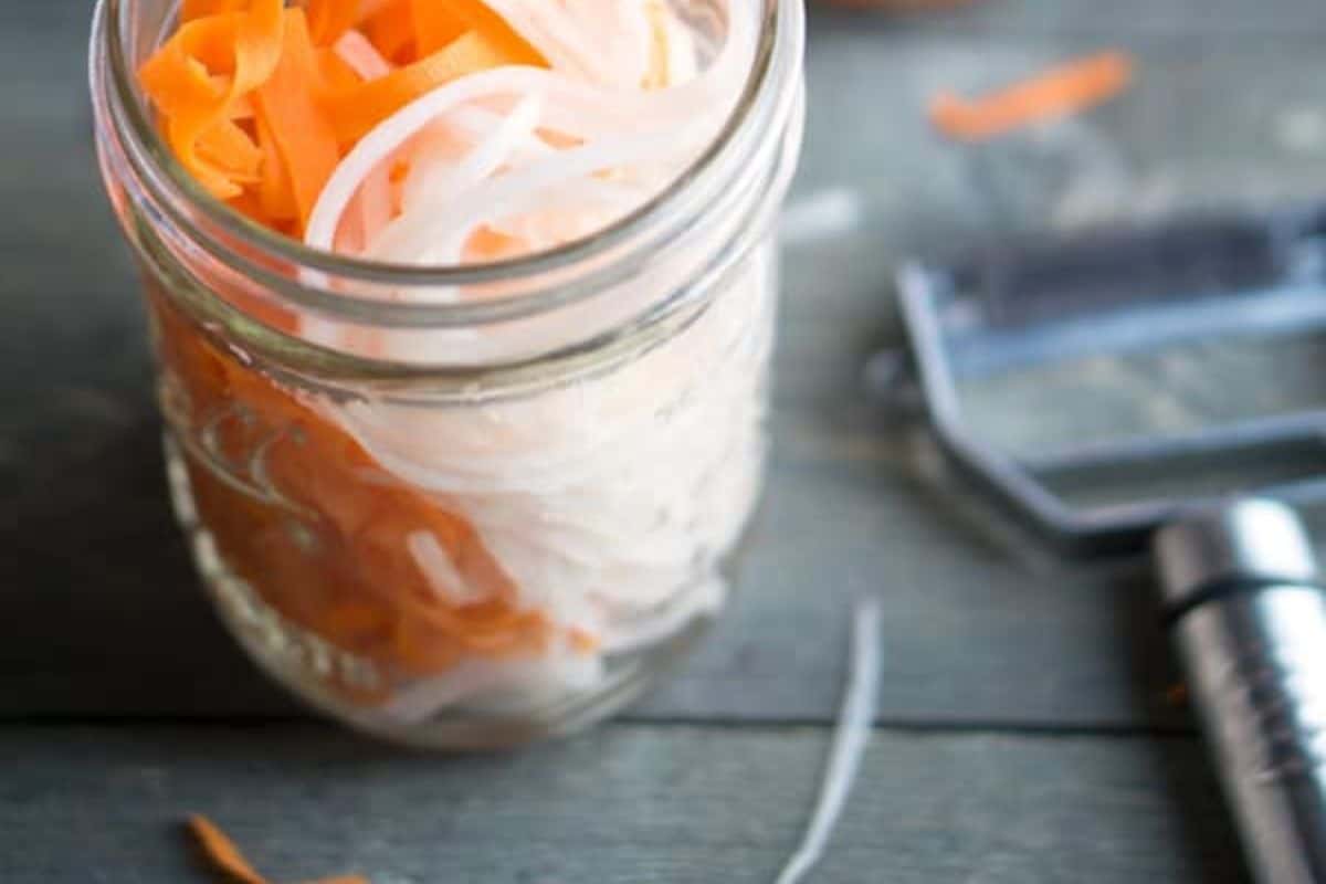 A clear glass mason jar filled with julienned orange carrots and white daikon radish, partially submerged in liquid. A vegetable peeler is visible in the background.