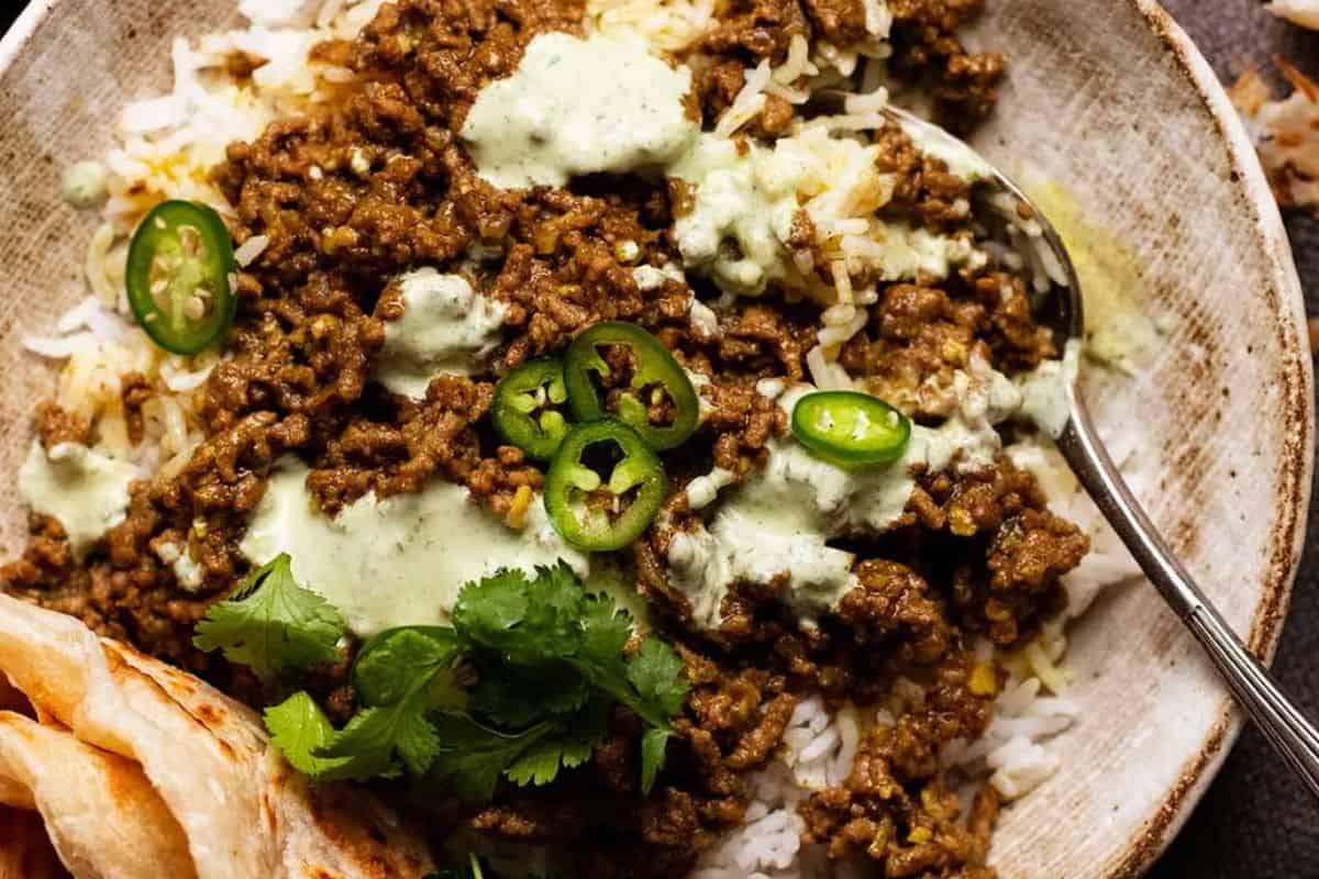 A bowl of white rice topped with ground meat in sauce, sliced green chili, fresh cilantro, and a drizzle of green herb sauce showcases bold flavors inspired by Indian recipes, served with a spoon and flatbread on the side.