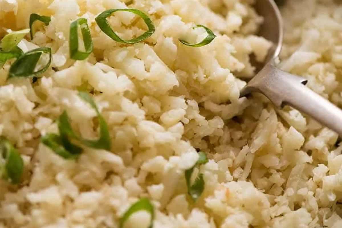 Close-up of cooked rice garnished with sliced green onions, showcasing a flavor packed dish, with a metal serving spoon partially visible on the right.