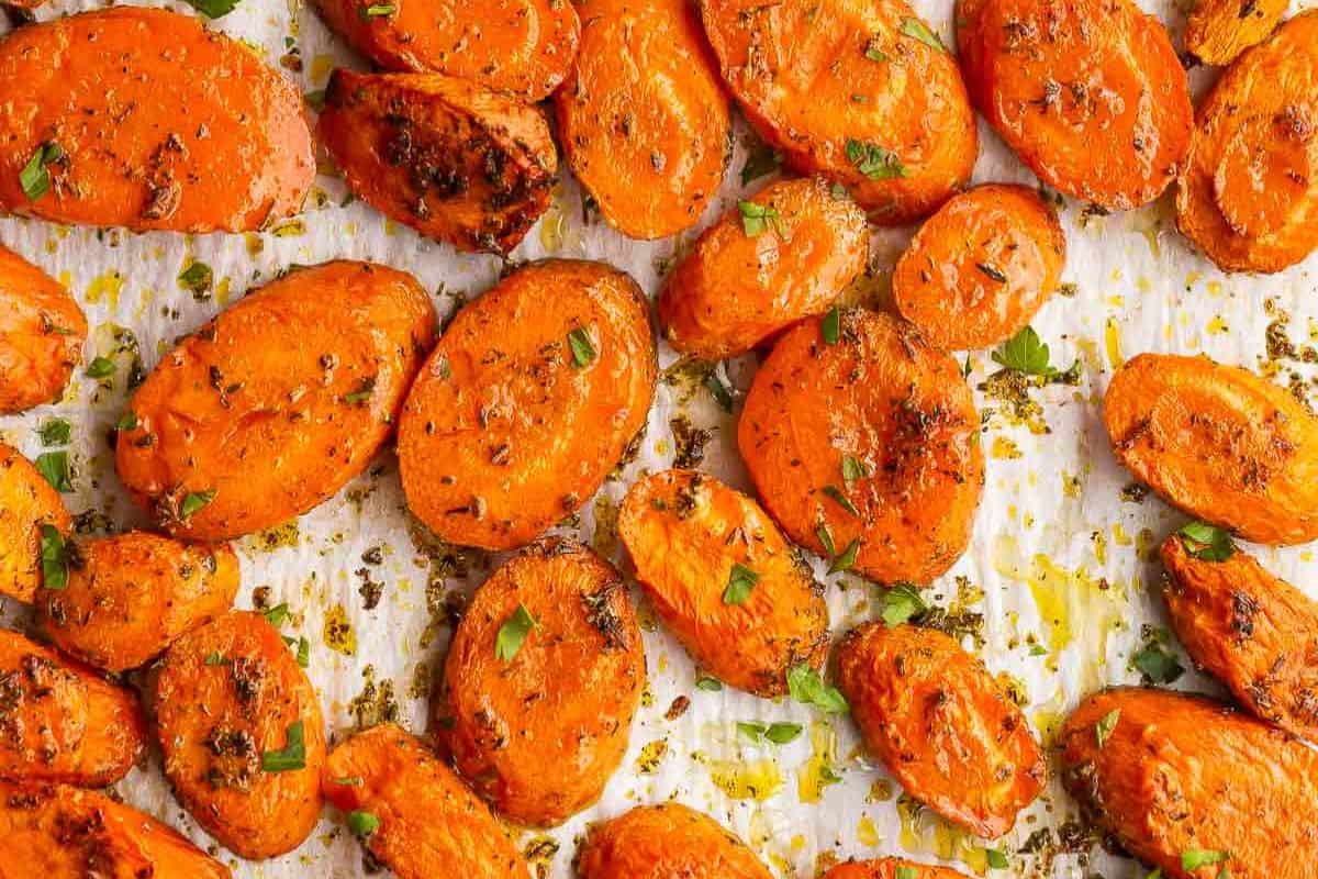 A close-up overhead shot of roasted carrot slices, seasoned with herbs and spices, scattered on a white sheet of parchment paper.