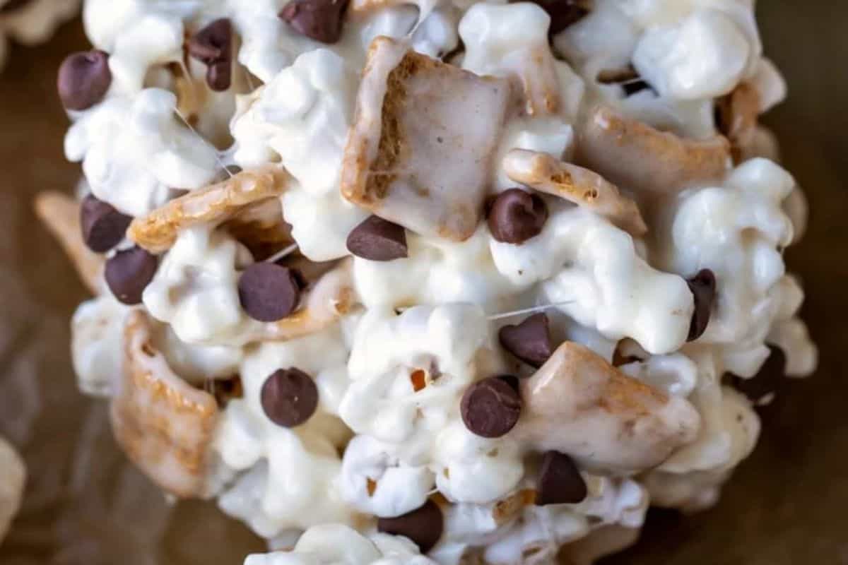 A close-up shot of a round, sticky "s'mores ball" made of popcorn, white coating (likely marshmallow), graham cracker pieces, and chocolate chips.