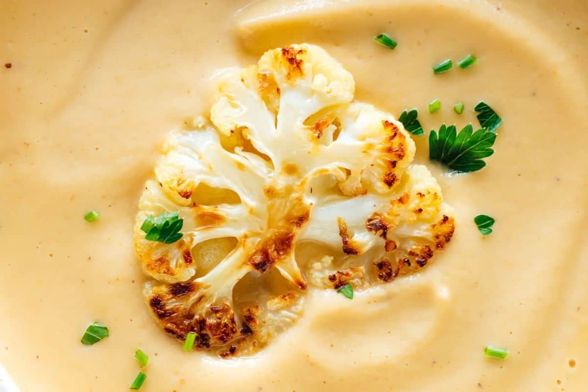 A bowl of creamy soup topped with a roasted cauliflower slice and garnished with chopped chives and parsley.