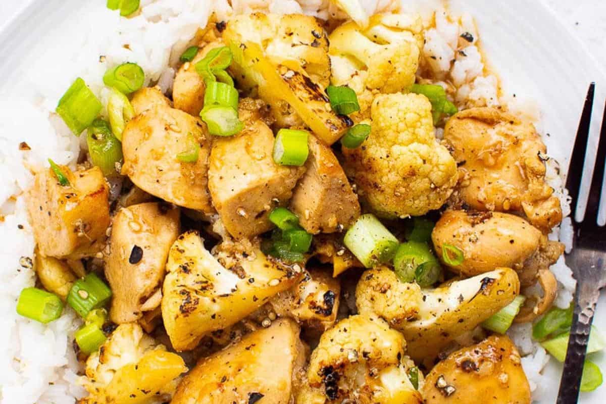 A plate of white rice topped with sautéed chicken, cauliflower florets, chopped green onions, and black pepper.
