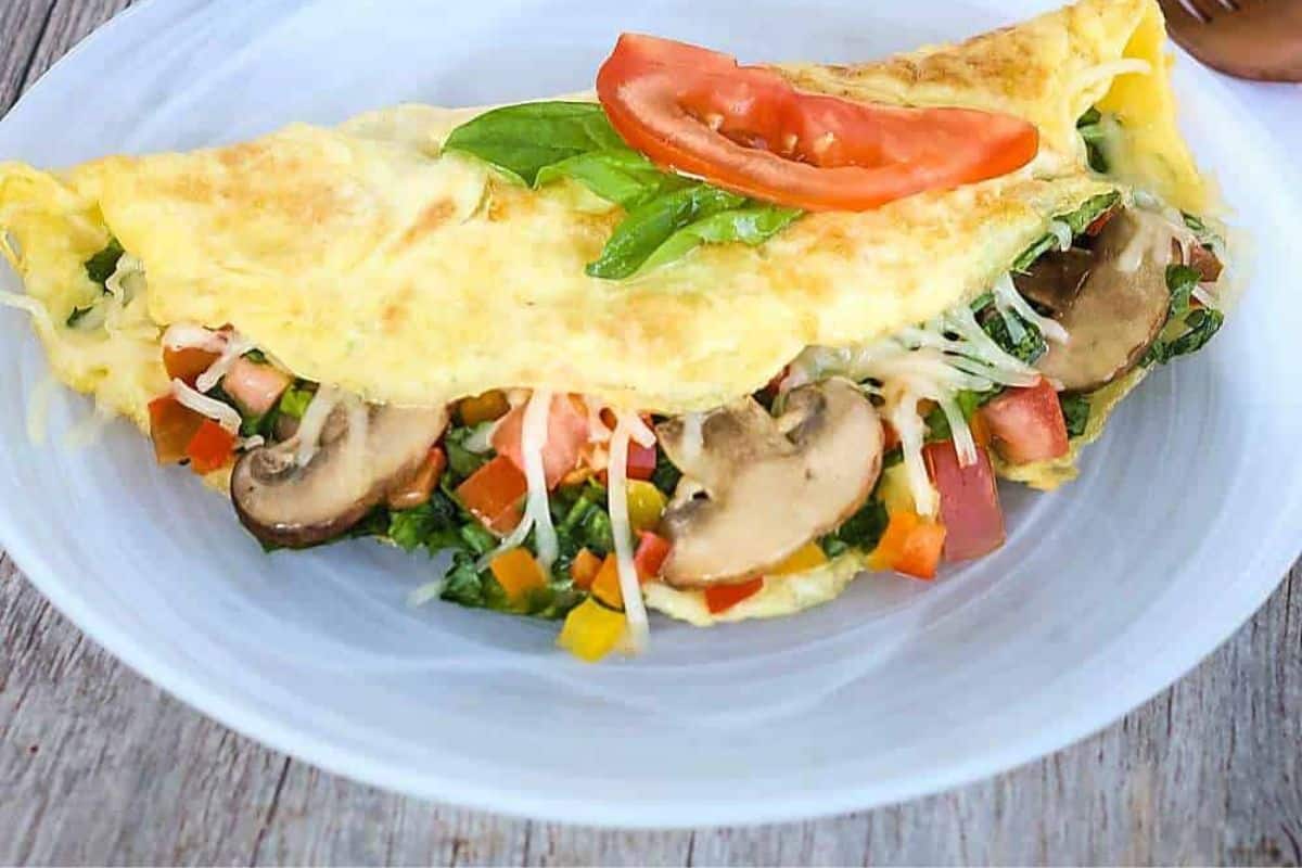 A close-up of an omelette with a slightly crispy outer layer, filled with sautéed spinach, diced bell peppers, onions, and mushrooms. Topped with a garnish and tomato