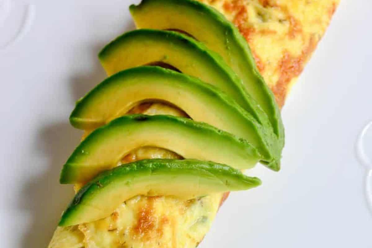 A golden-brown omelet with a slightly crispy exterior, topped with fresh avocado slices arranged neatly. The dish is served on a plate, highlighting the vibrant green of the avocado against the warm tones of the omelet. A simple yet visually appealing meal.