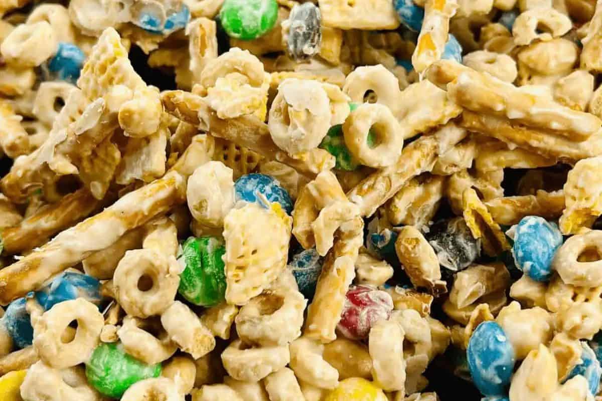 Close-up of white trash candy snack mix featuring pretzel sticks, Cheerios, Chex cereal, M&M candies, and other ingredients coated in a white chocolate mixture.
