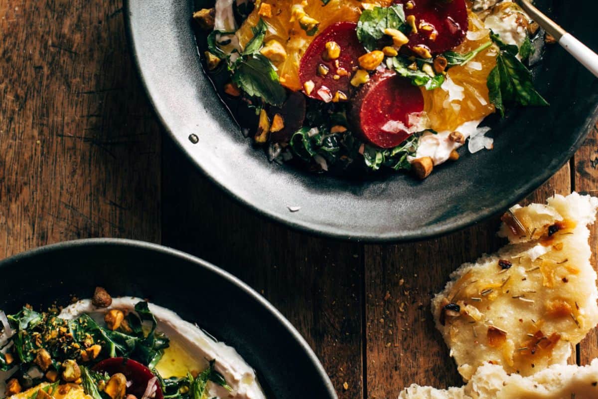 A top-down view of two dark bowls containing a vibrant salad with sliced beets, orange segments, green leafy vegetables, and chopped pistachios, resting on a rustic wooden table. A piece of focaccia bread is in the foreground.