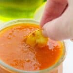 A hand dipping a piece of Air Fryer Gnocchi into a glass bowl filled with orange dipping sauce. A blurred glass with a yellow-green liquid is visible in the background.
