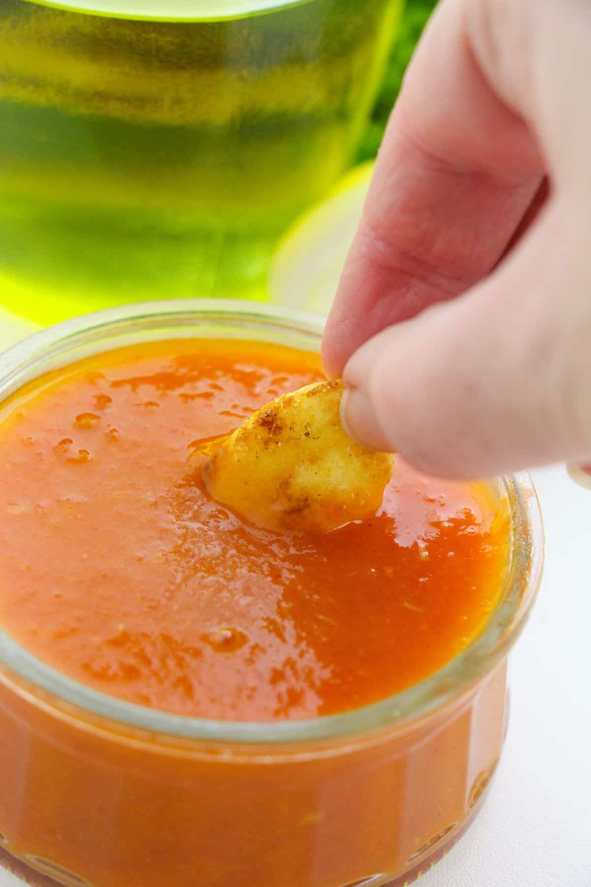 A hand dipping a piece of Air Fryer Gnocchi into a glass bowl filled with orange dipping sauce. A blurred glass with a yellow-green liquid is visible in the background.