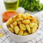 A white bowl filled with Air Fryer Gnocchi sits on a checkered cloth, with a jar of red sauce, a bottle of oil, fresh parsley, and a garlic bulb in the background.