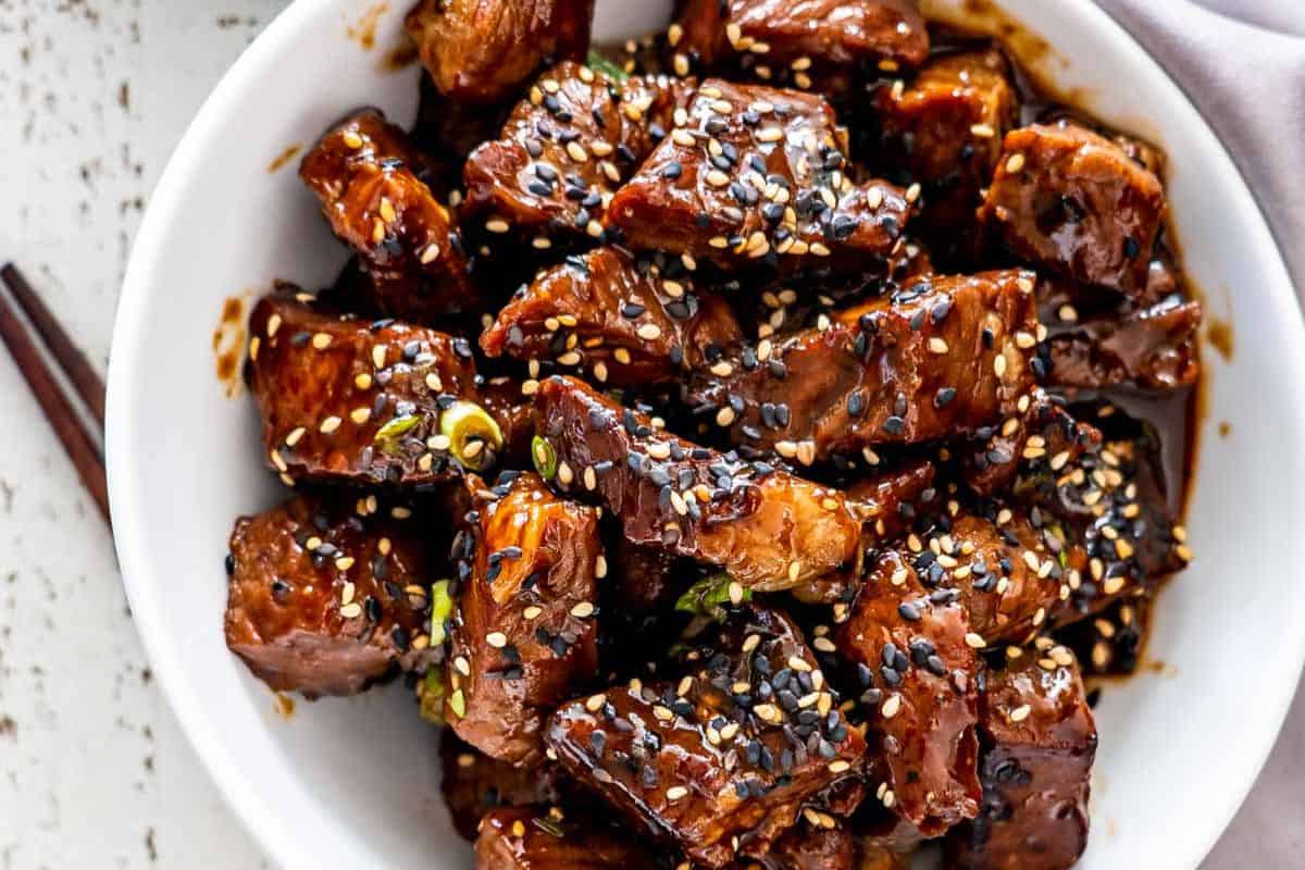 A bowl of glazed beef cubes topped with black and white sesame seeds, perfect for fans of juicy steak and classic steakhouse flavors.