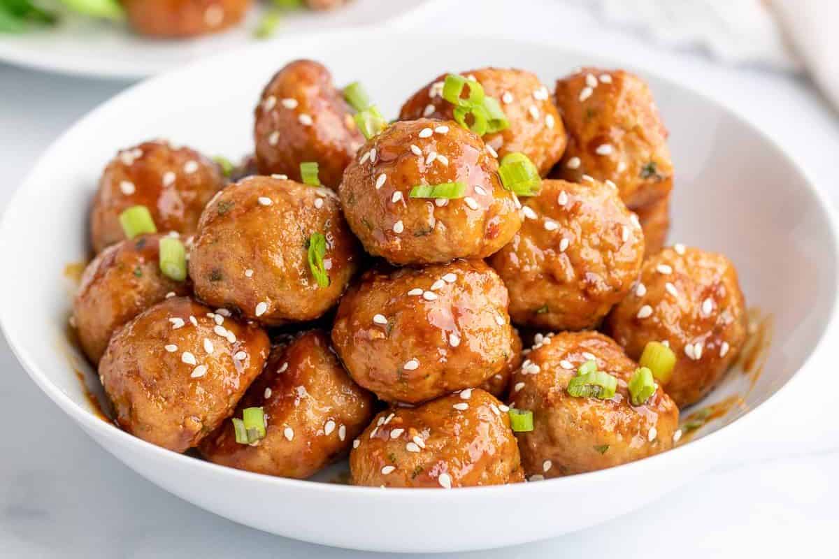 A white bowl filled with glazed meatballs, topped with sesame seeds and chopped green onions—perfect for those seeking cold appetizer ideas.