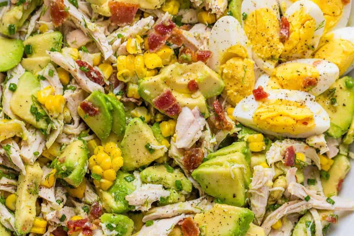 Close-up of a salad with avocado, corn, canned chicken, chopped bacon, and sliced hard-boiled eggs, seasoned with black pepper—perfect for easy dinner recipes and budget friendly dinners.