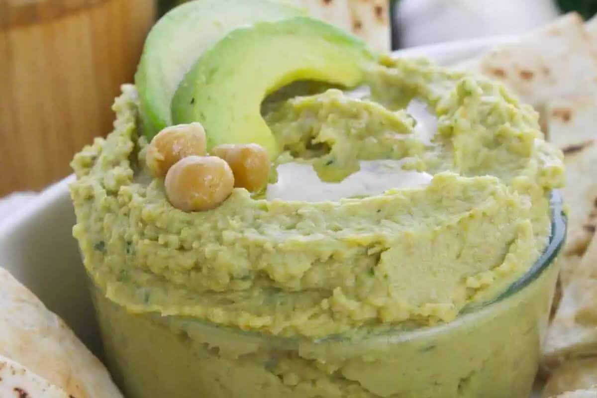 A glass bowl filled with green avocado hummus—one of the most delicious twists on classic hummus—topped with chickpeas and avocado slices, surrounded by pieces of flatbread.