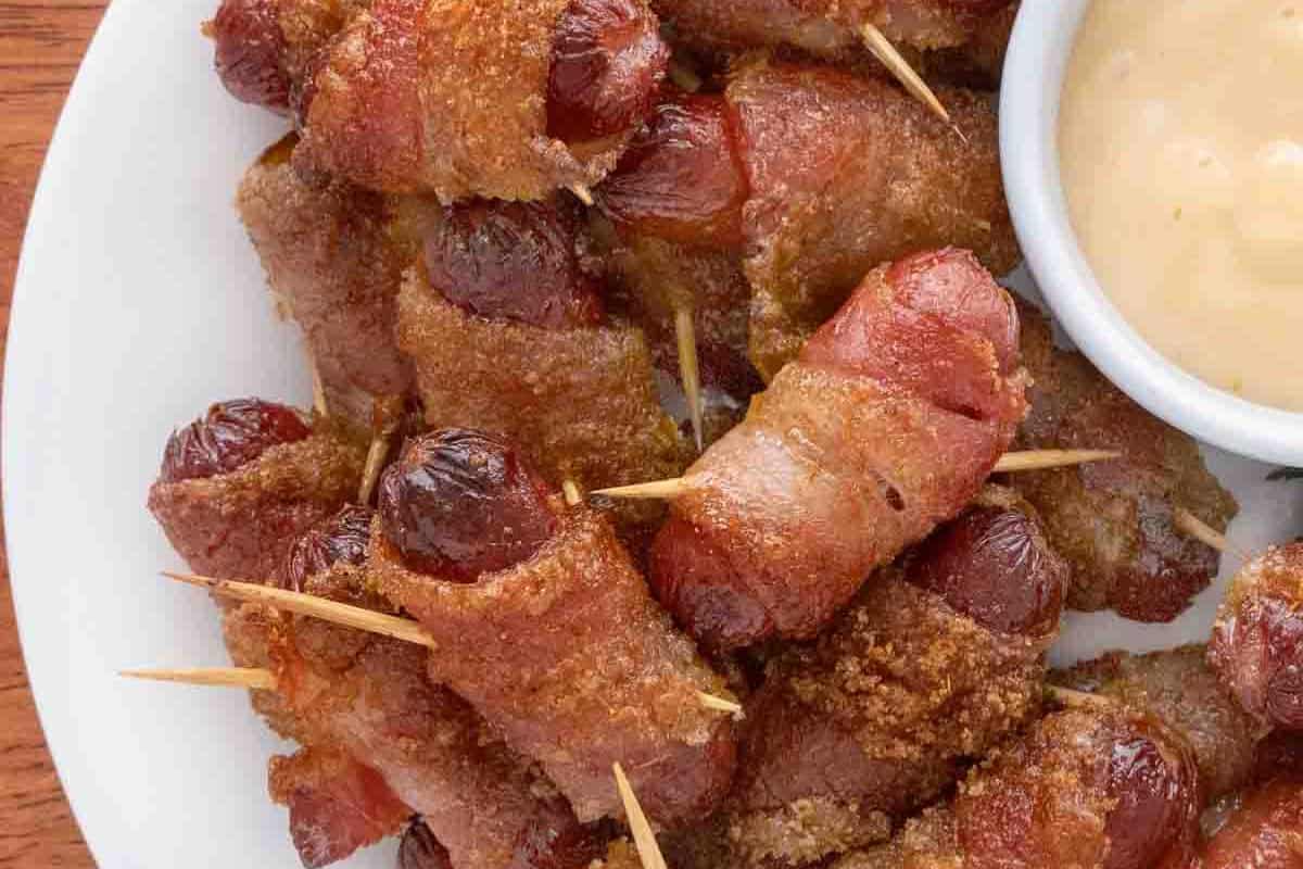 Bacon-wrapped sausages secured with toothpicks on a plate, served with a side of dipping sauce—perfect for those seeking tasty cold appetizer ideas.
