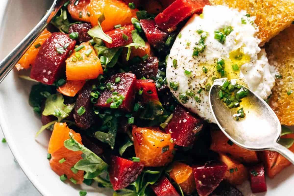  A close-up of a beet and burrata salad featuring chunks of red and orange beets, orange segments, arugula, and a large dollop of creamy burrata cheese drizzled with olive oil and chives. Toasted bread is visible on the side